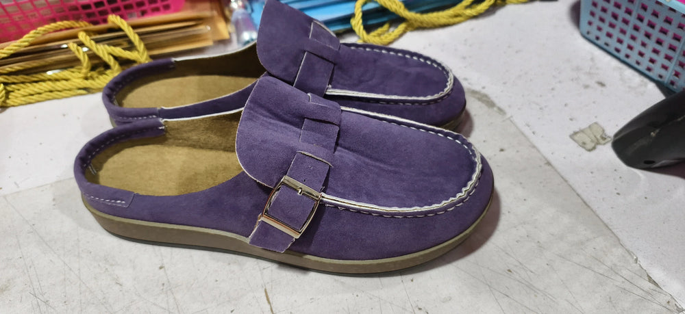 Pair of purple suede slip-on loafers with buckle detail and beige soles on a white surface