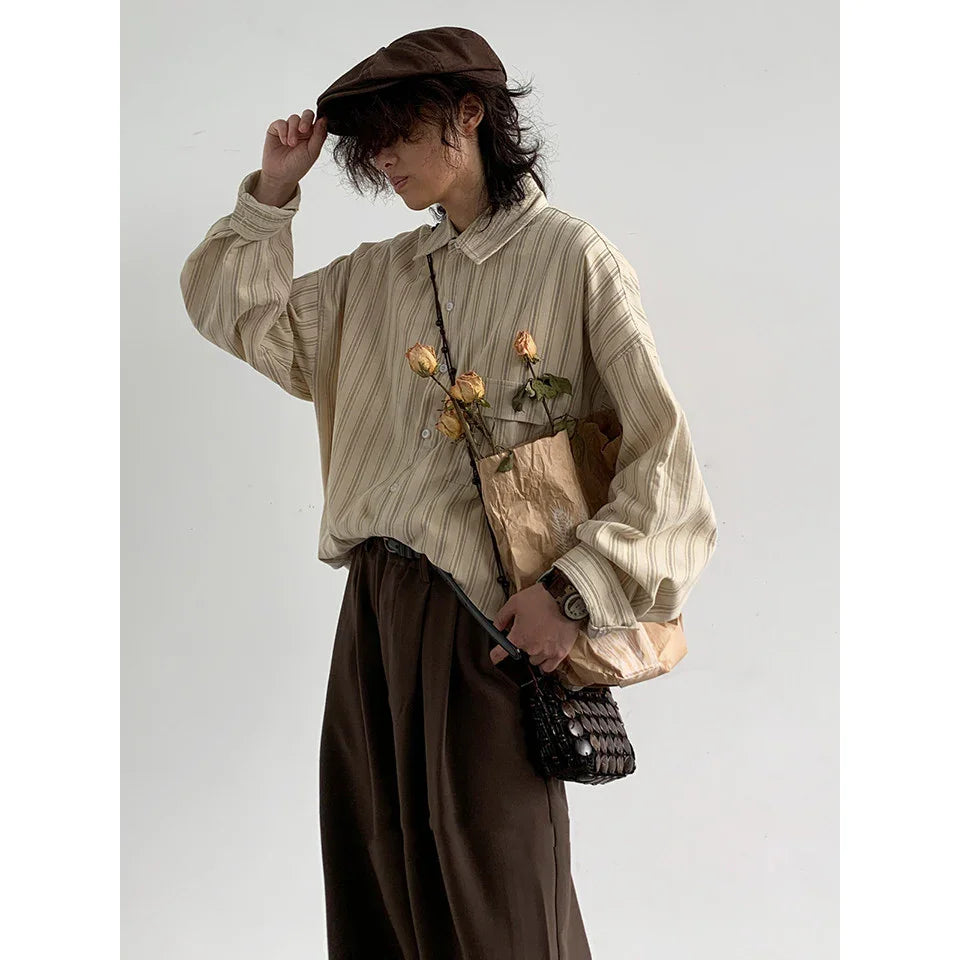 Person wearing beige striped oversized shirt, dark brown pants, and brown cap holding bag with dried roses