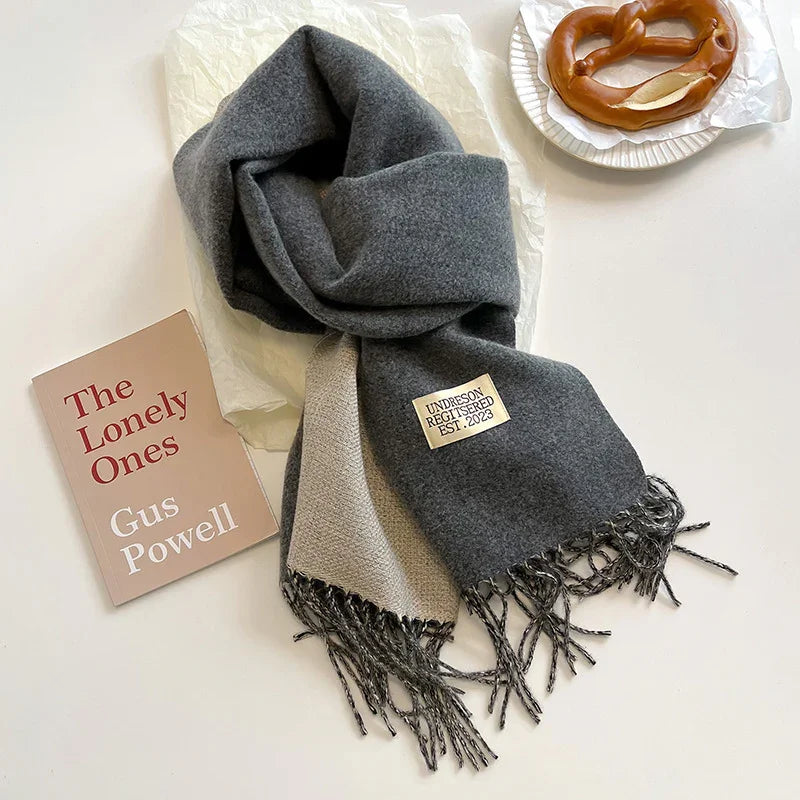 Grey and beige fringed wool scarf with label, book titled The Lonely Ones by Gus Powell, and soft pretzel on plate