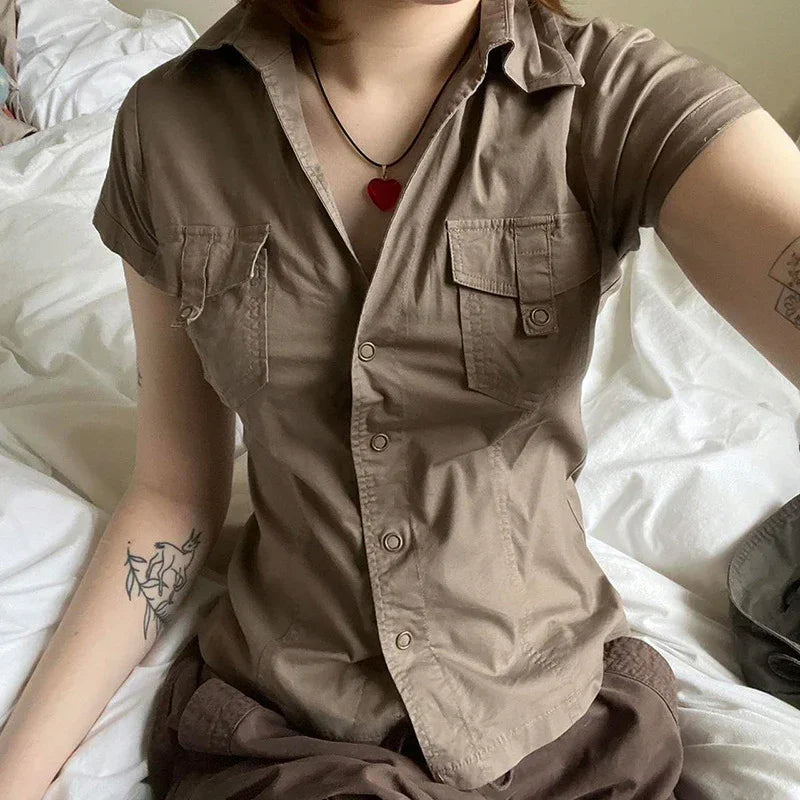 Person wearing brown short-sleeve button-up shirt with chest pockets and red heart necklace sitting on white bed