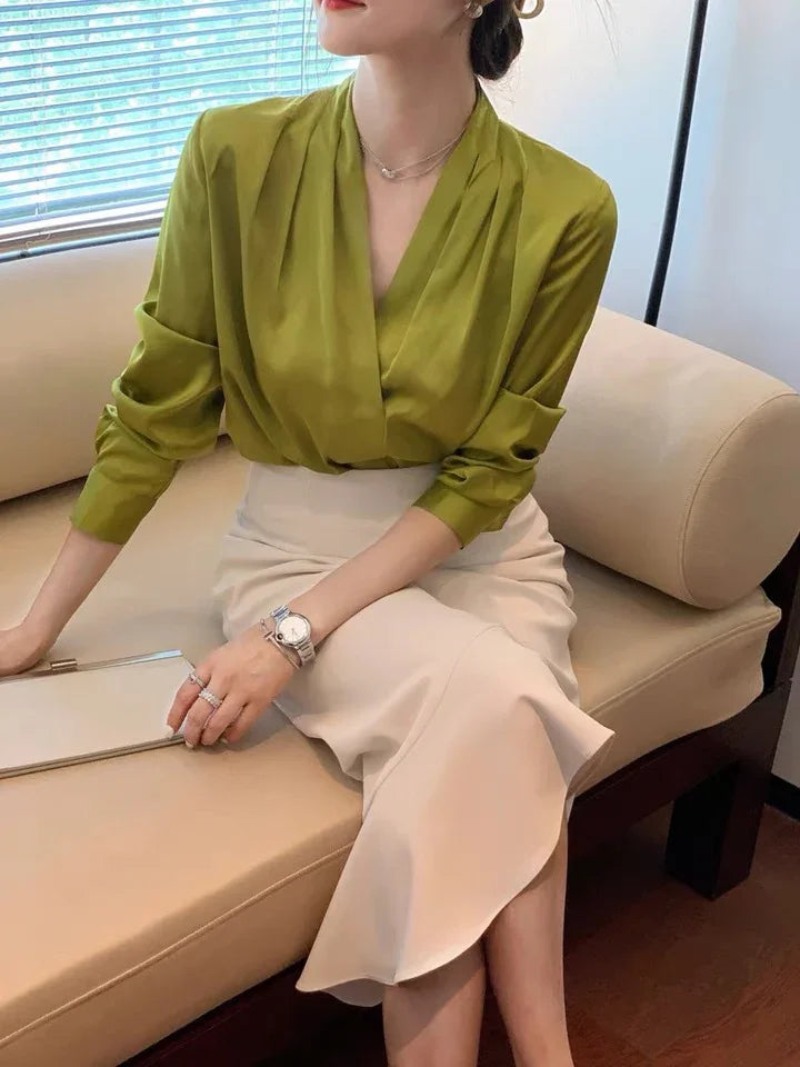 Woman seated on beige couch wearing olive green blouse and cream midi skirt, accessorized with silver watch and rings