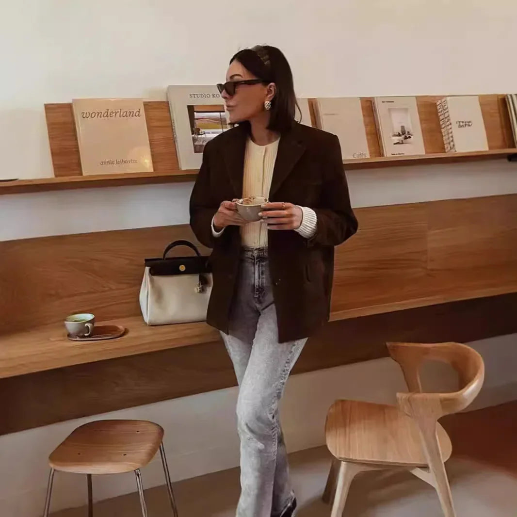 Woman in brown blazer and jeans holding coffee in modern cafe with wooden furniture and art books