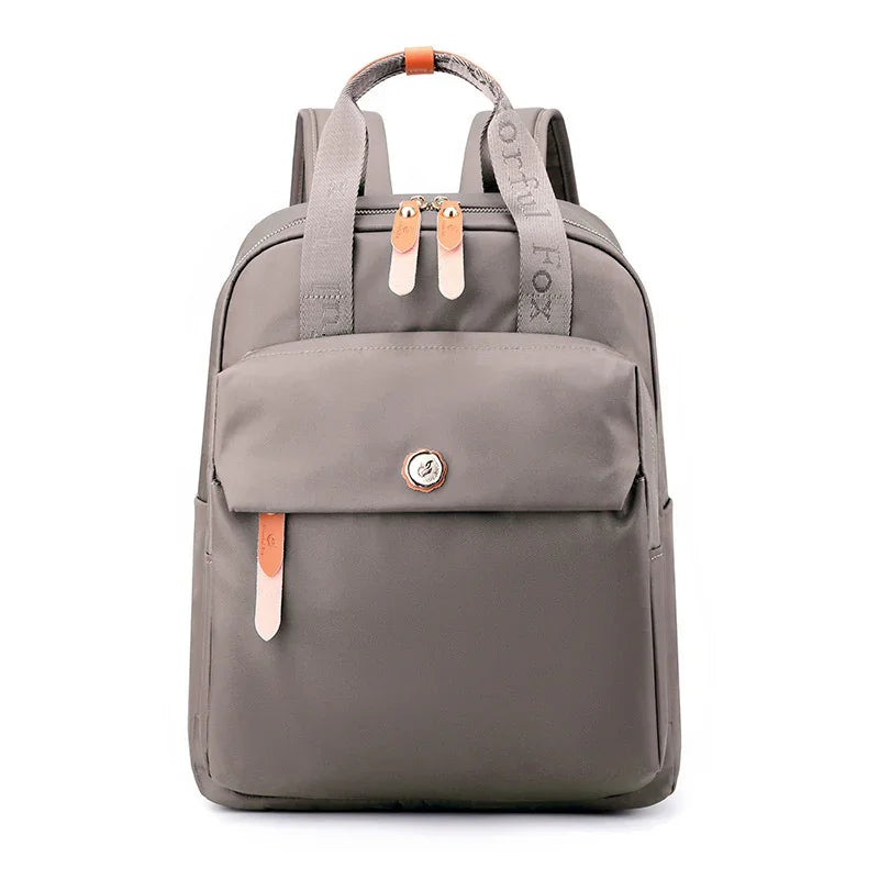 gray compact backpack with front pocket, dual zippers, leather accents, and branded fabric handle