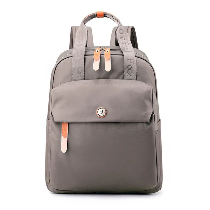 gray compact backpack with front pocket, dual zippers, leather accents, and branded fabric handle