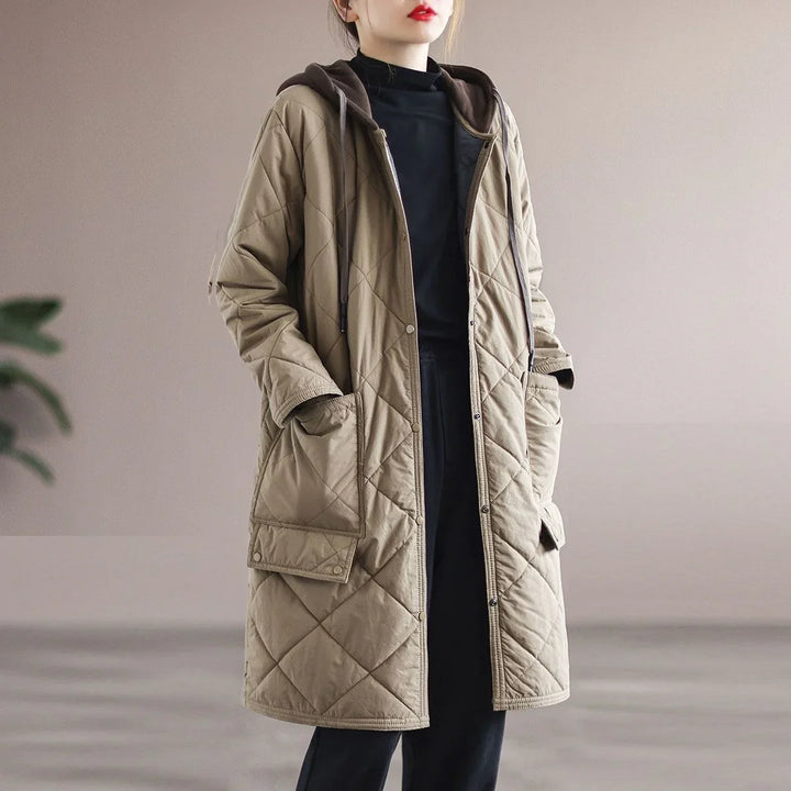 Woman wearing long beige quilted coat with hood and large pockets over black outfit indoors