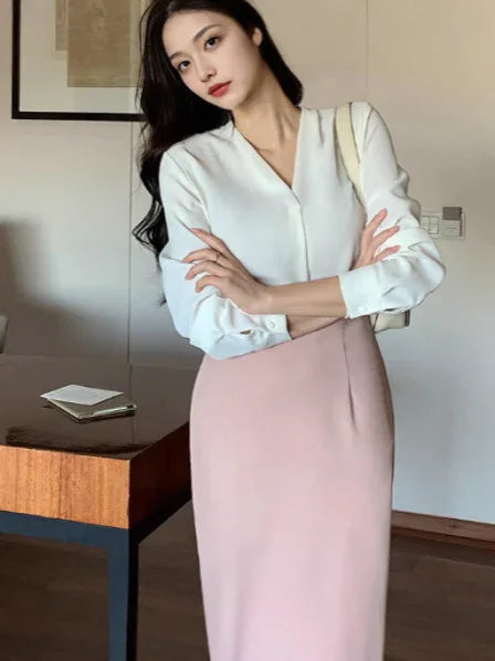 Woman in white blouse and pastel pink skirt standing indoors next to a wooden table