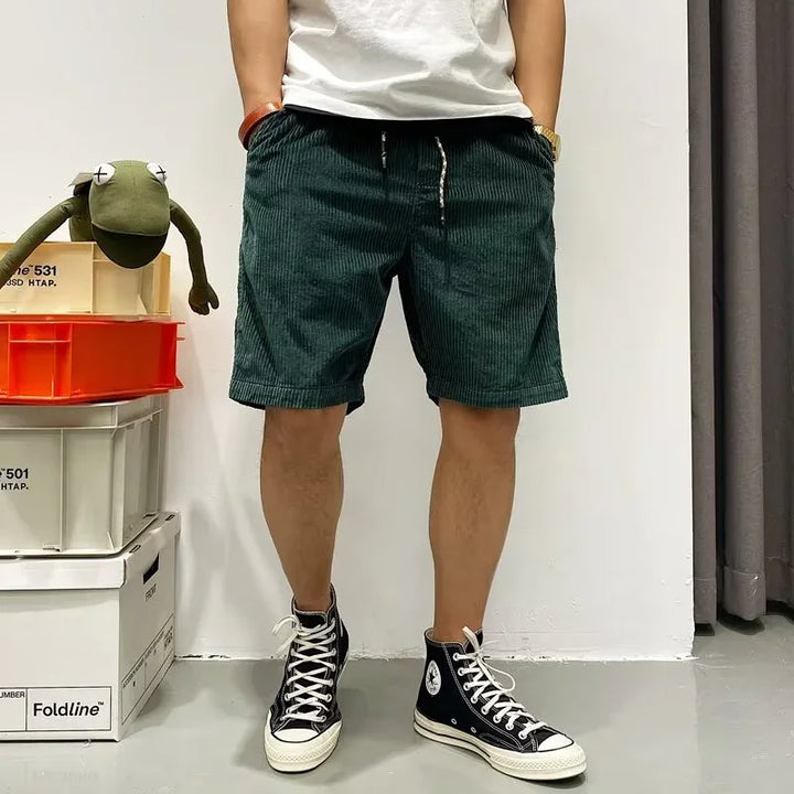 Man wearing dark green corduroy shorts and black high-top sneakers standing indoors next to stacked storage boxes