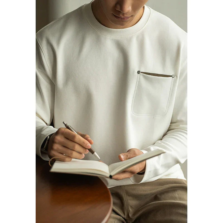 Man wearing white pocketed sweatshirt writing in open notebook at wooden table