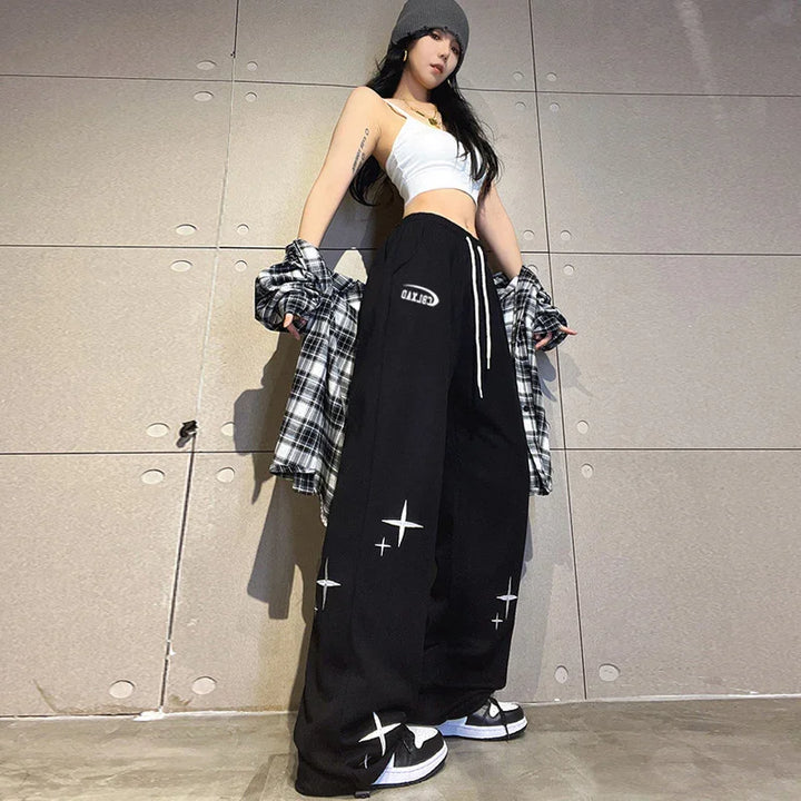 young woman wearing black wide-leg pants with white star designs, white crop top, gray beanie, and checkered jacket