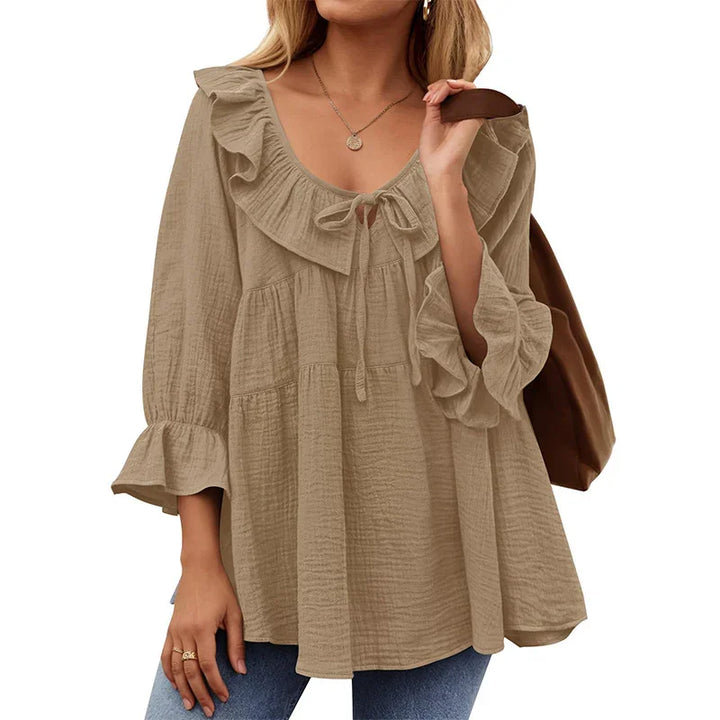 Woman wearing beige ruffled long sleeve blouse with scoop neck and bow detail, paired with blue jeans