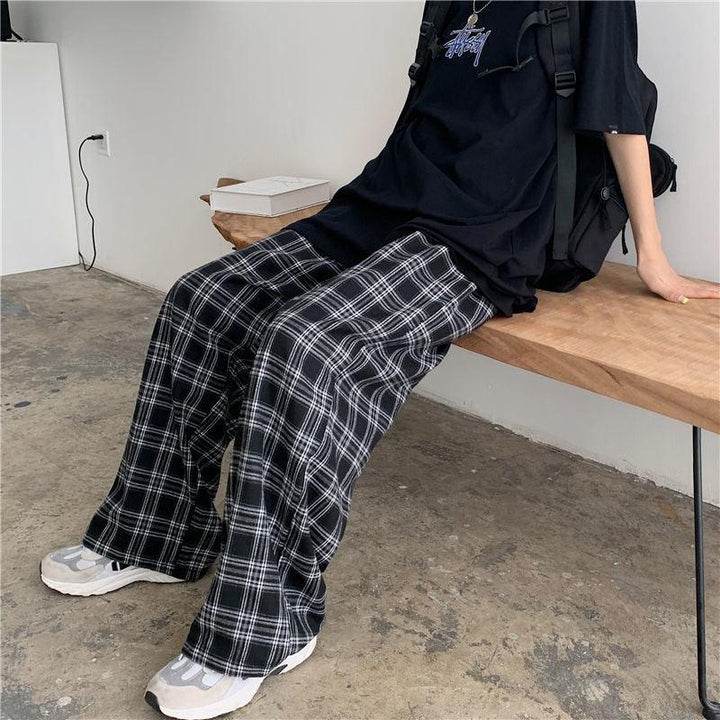 Person wearing black plaid wide-leg pants, black graphic t-shirt, white sneakers, sitting on wooden bench in minimalist room