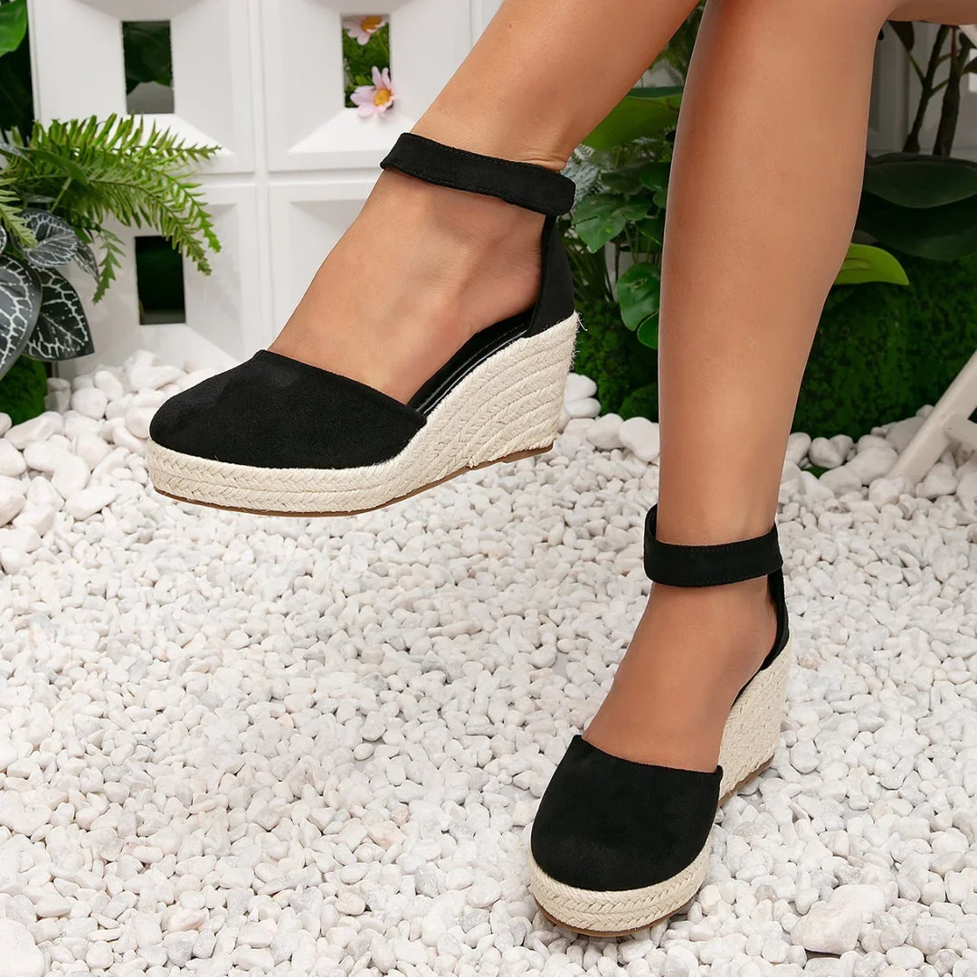Black suede wedge espadrille sandals with ankle straps on feet, standing on white stones with green plants background