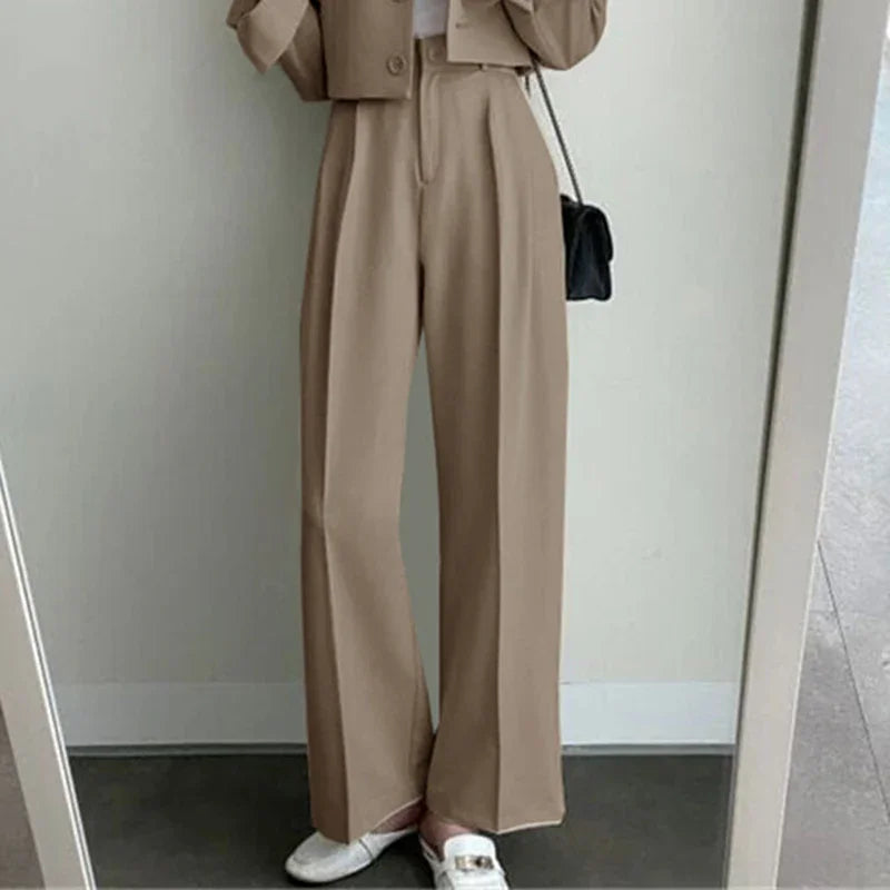 Women wearing beige wide-leg trousers with white loafers and black shoulder bag in minimal indoor setting