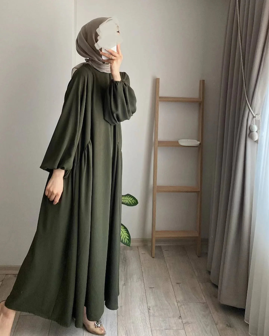 Woman wearing olive green modest maxi dress and beige hijab taking a mirror selfie in a minimal room