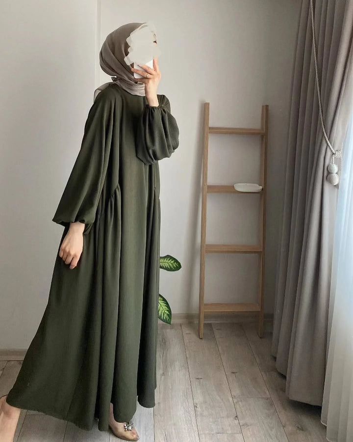 Woman wearing olive green modest maxi dress and beige hijab taking a mirror selfie in a minimal room