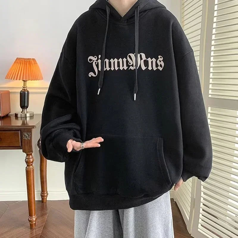 Person wearing oversized black hoodie with gothic font text, grey pants in cozy indoor setting