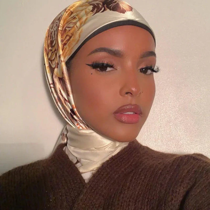 Close-up portrait of woman wearing floral satin hijab and brown knit sweater against plain wall