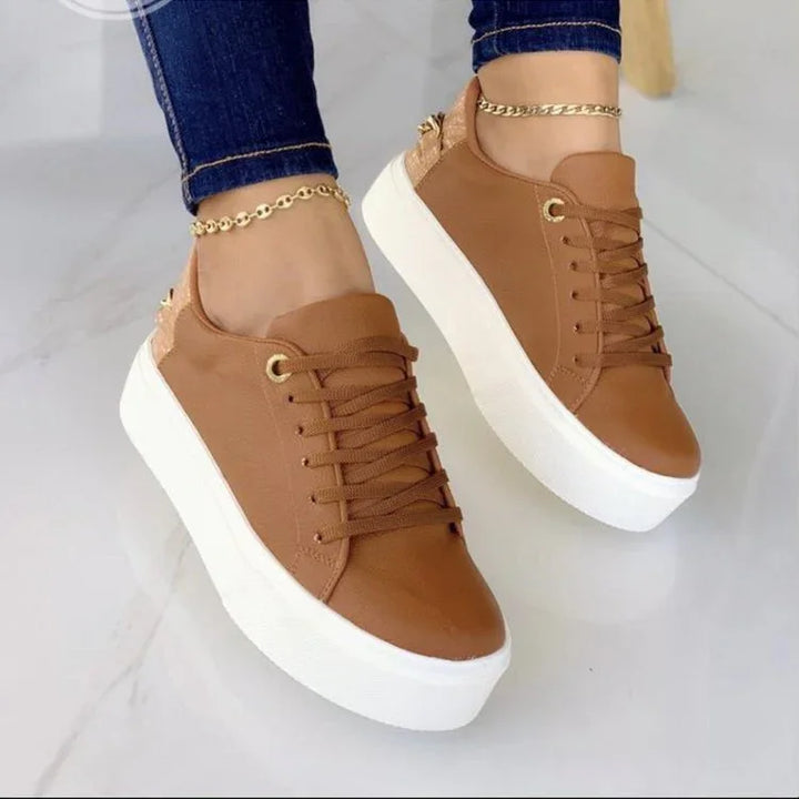 Brown leather platform sneakers with thick white soles worn with blue jeans and gold anklets on white marble floor