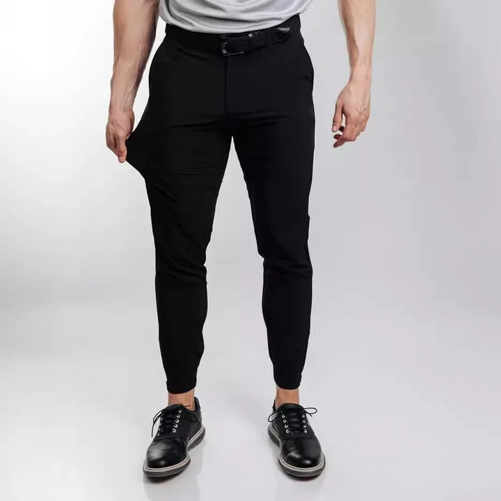 Man wearing slim-fit black stretch pants pulling fabric to show flexibility, paired with black dress shoes