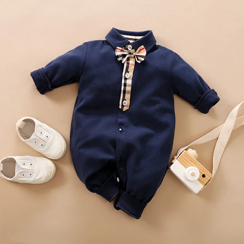 Navy blue baby romper with plaid bow tie and buttons, paired with white baby shoes and a wooden toy camera