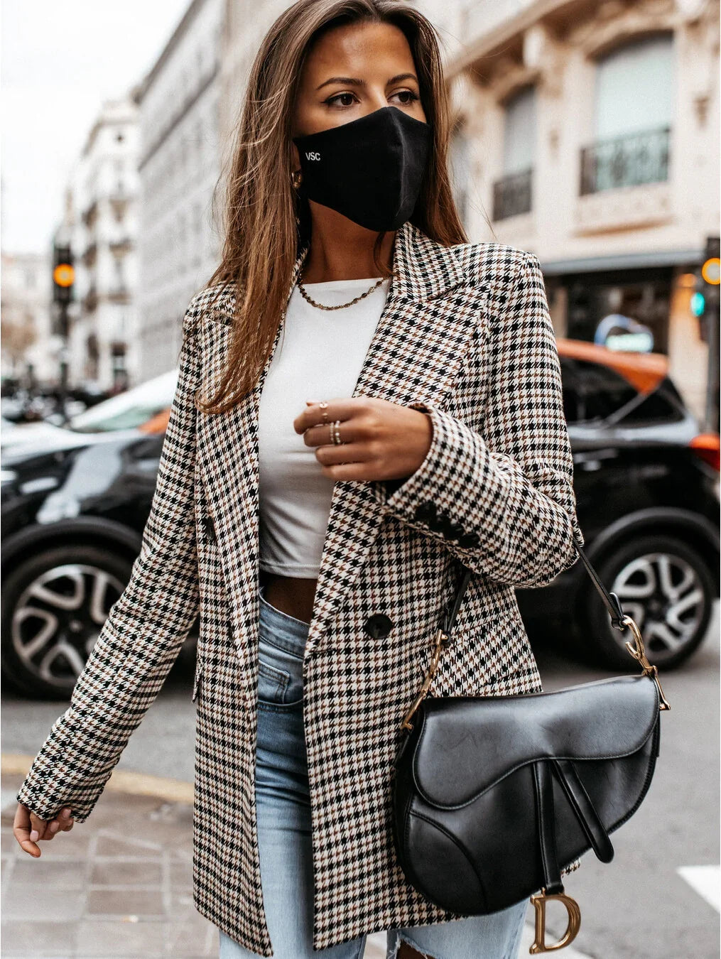 Stylish woman in houndstooth blazer, white crop top, denim jeans, black face mask, and black leather handbag on city street