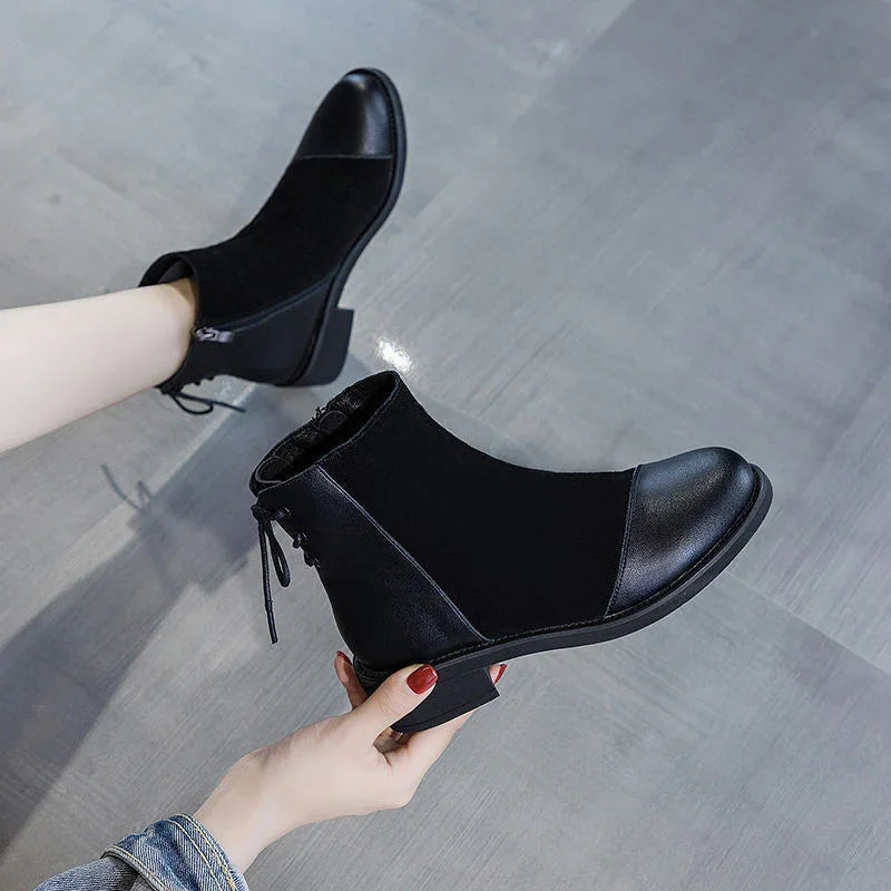 Black ankle boots with leather toe caps and low heels on gray floor, worn by person with red nail polish