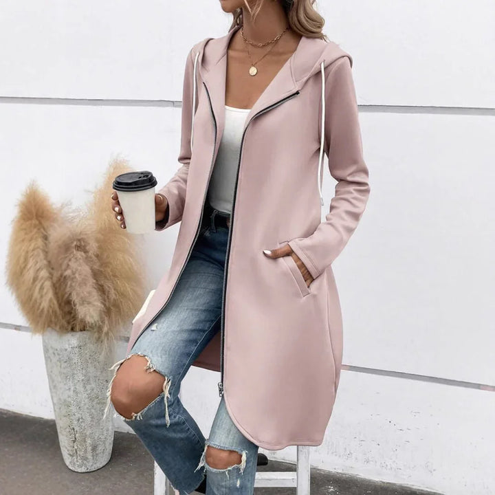 Woman wearing light pink hooded zip-up long jacket with pockets and ripped blue jeans holding coffee cup