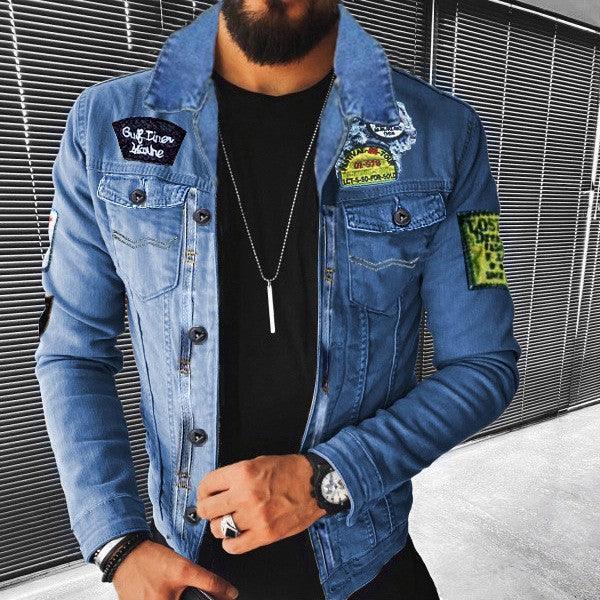 Man wearing blue denim jacket with patches over black shirt, accessorized with necklace and watch