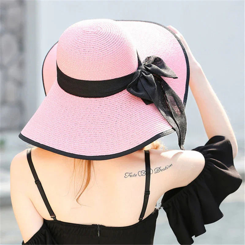 Woman wearing a large pink sun hat with black trim and bow, black off-shoulder top, tattoo visible on upper back