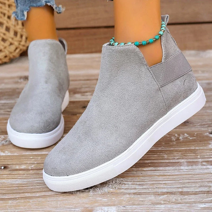 Women's grey suede slip-on sneakers with white soles on wooden floor, styled with turquoise beaded anklet