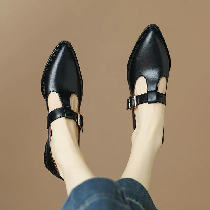 Black leather pointed toe flats with buckle straps worn with blue jeans on beige background