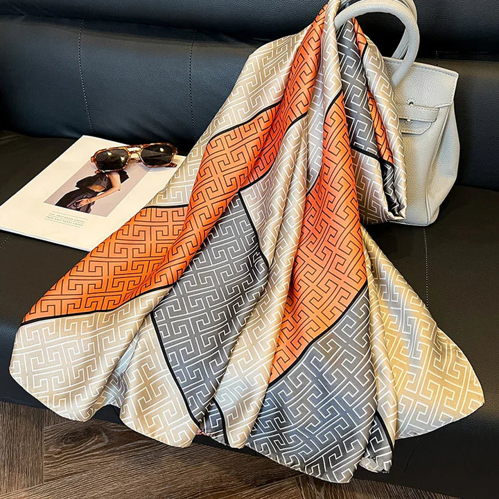 Silk scarf with orange, beige, and gray geometric pattern draped over beige handbag on black leather couch with sunglasses and fashion magazine