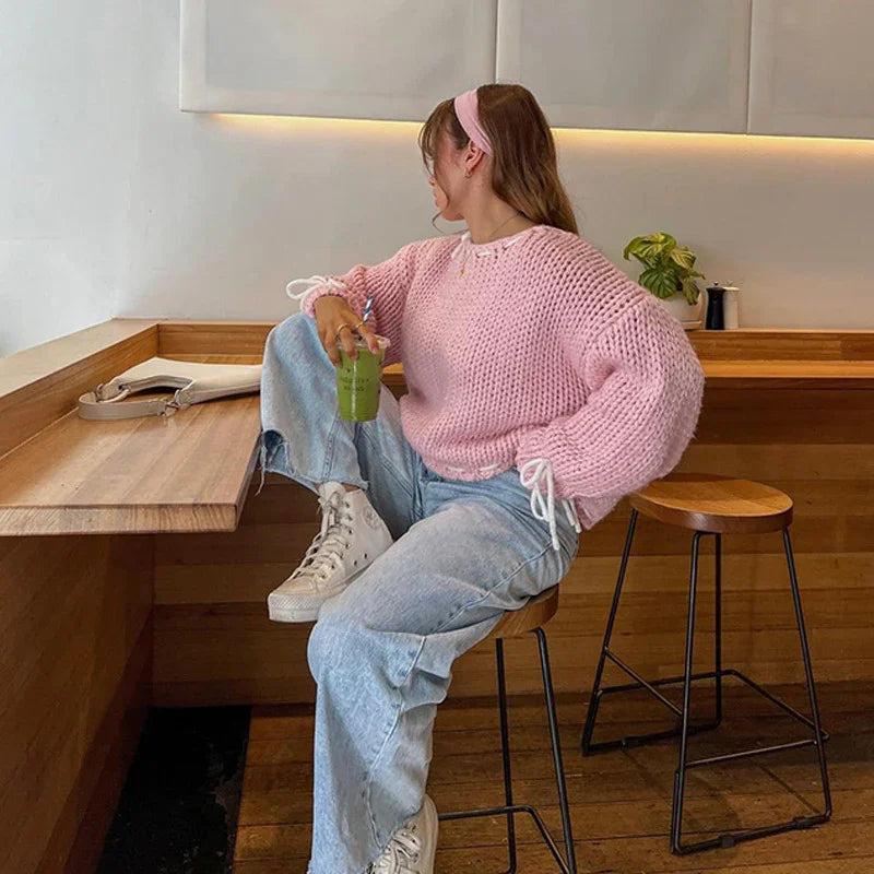 Woman in pink chunky knit sweater and light blue jeans sitting on wooden stool, holding green smoothie in modern cafe