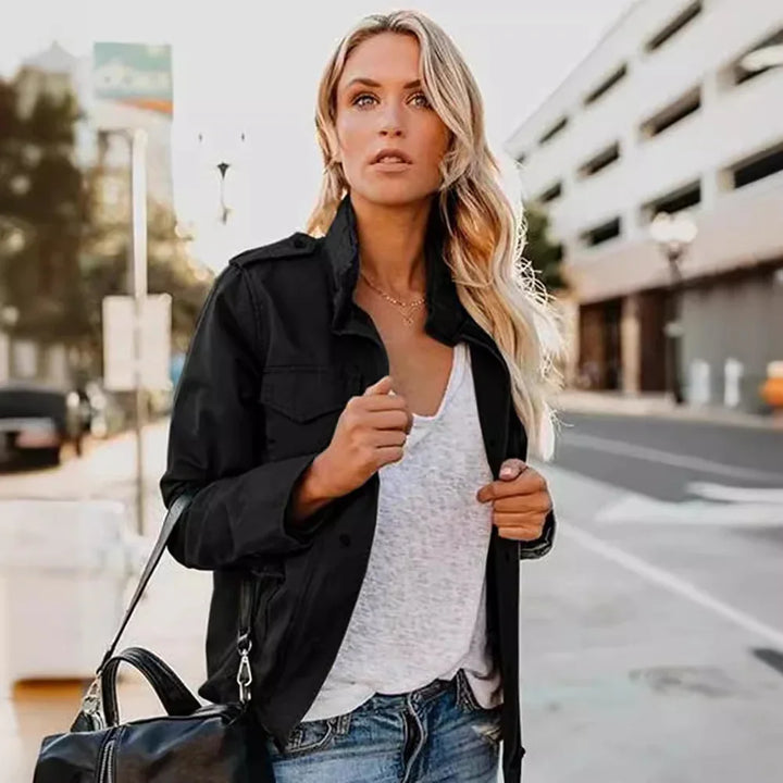 Blonde woman wearing a black jacket, white shirt, and jeans posing outdoors with a black bag