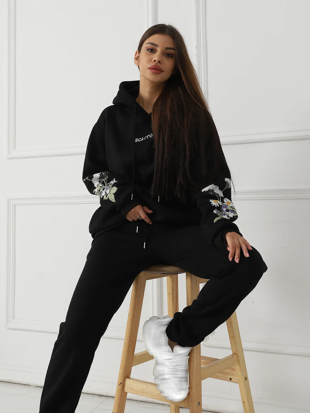Woman in black hoodie with floral sleeves and black pants sitting on wooden stool with white sneakers