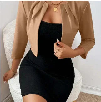 Woman wearing fitted black dress with camel-colored cropped blazer, gold layered necklaces, seated on white chair