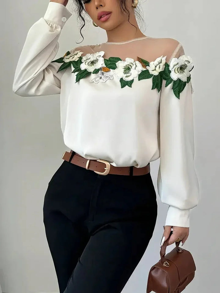 Woman wearing white blouse with floral embroidery and sheer neckline, black pants, brown belt, holding brown handbag