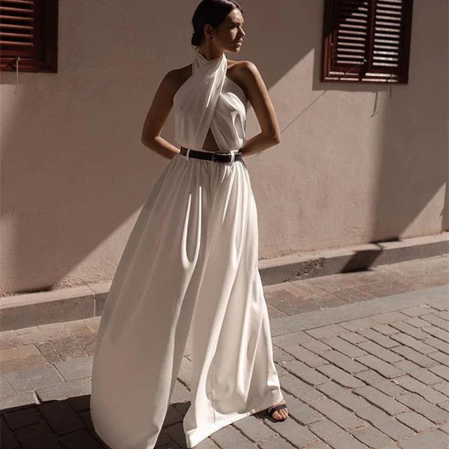 Woman wearing elegant white halter neck dress with black belt posing on sunlit street with shuttered windows