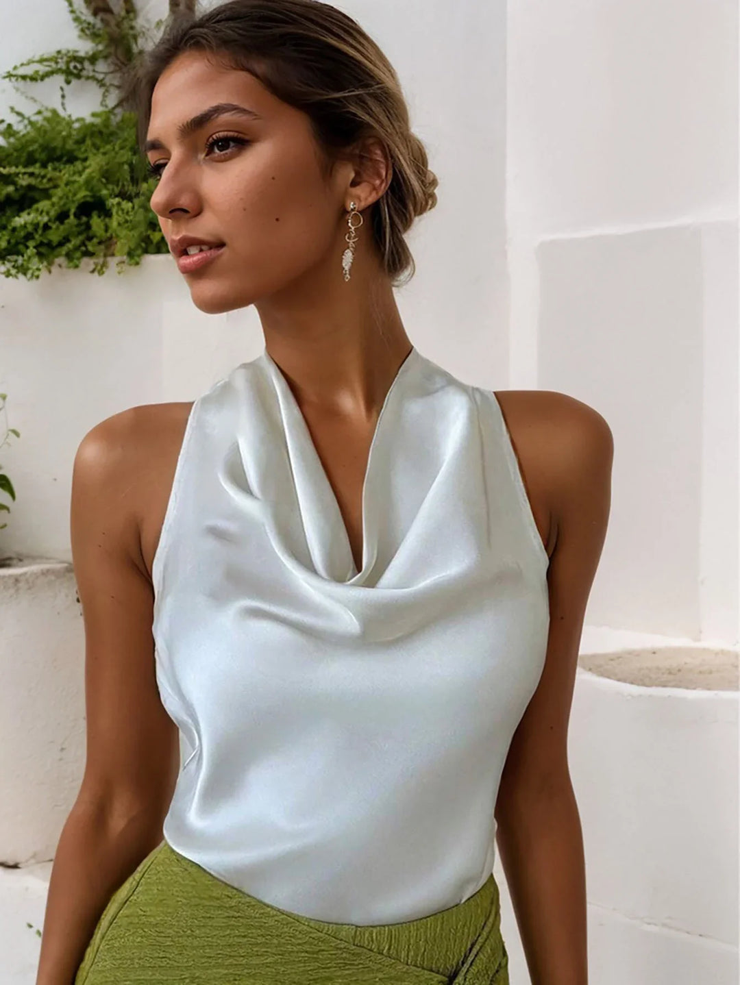 Woman wearing light blue satin halter top and green textured skirt with gold drop earrings against white wall