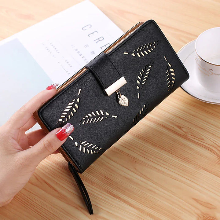 Black leather wallet with cut-out leaf designs and gold leaf charm held by hand on wooden table