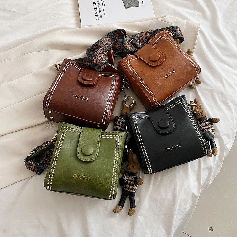 Four square leather handbags in brown, tan, green, and black with button closures and decorative stitching on white fabric