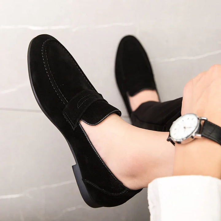 Black suede men's loafers worn with black pants and a silver wristwatch on a light floor