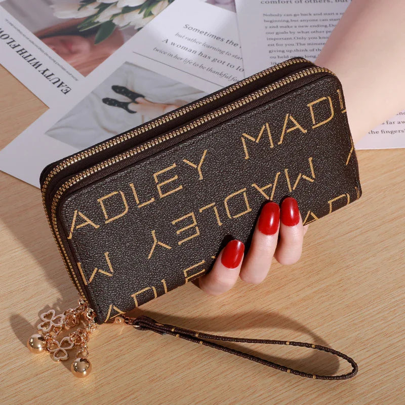 Brown zippered wristlet wallet with gold lettering and charm, held by hand with red nail polish