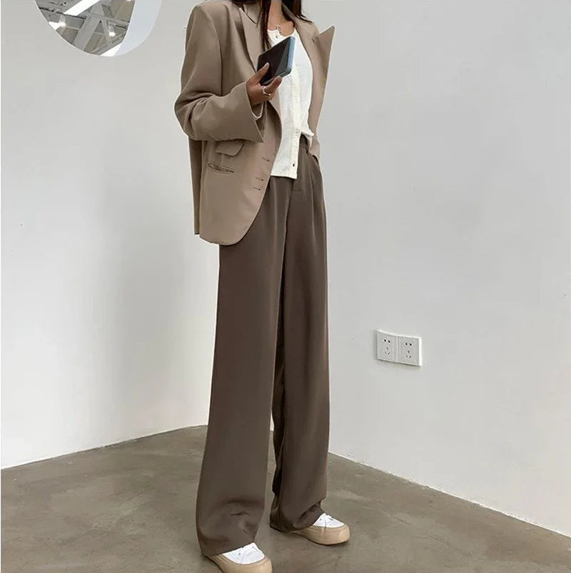 Woman wearing beige blazer, white top, wide brown pants, and white sneakers holding phone indoors