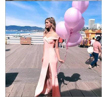 Woman in pink off-shoulder slit dress holding pink balloons standing on sunny beach boardwalk