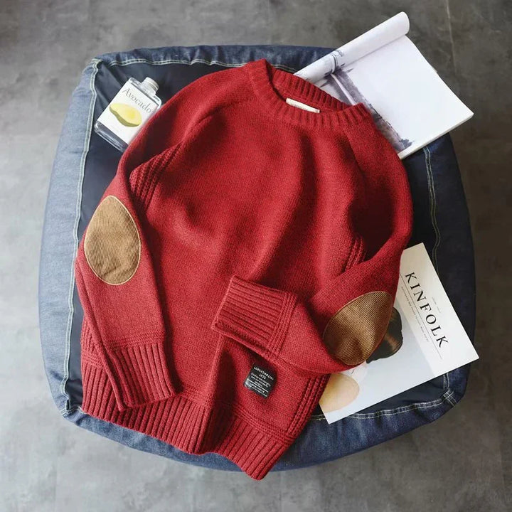 Red knitted sweater with brown elbow patches laid on a blue cushion with a magazine and an avocado bottle