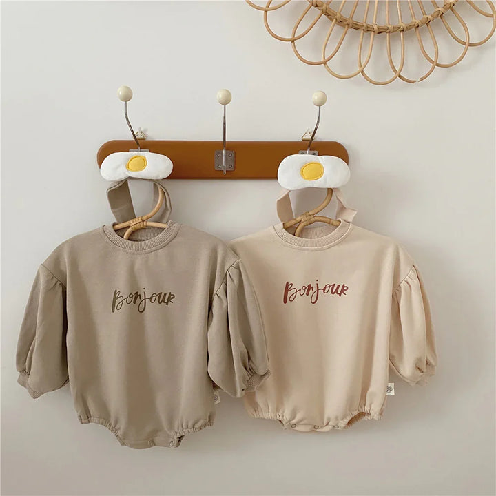 Two beige baby rompers with puff sleeves and the word 'Bonjour' hanging on hooks with fried egg hat accessories