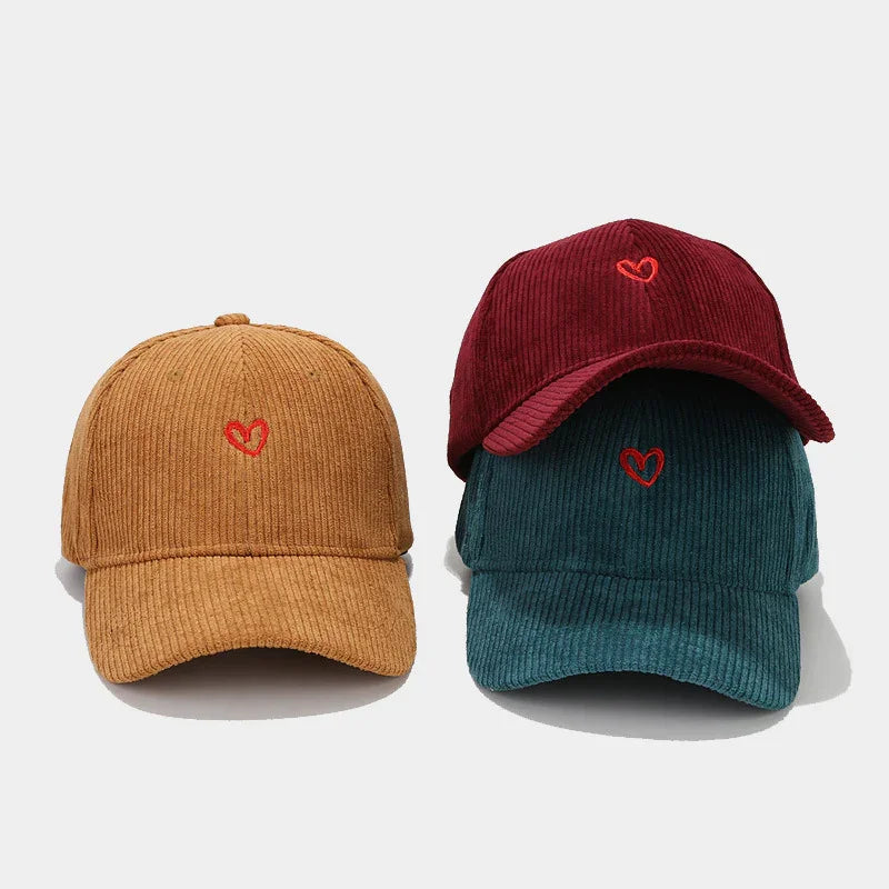 Three corduroy baseball caps in tan, burgundy, and teal with small red heart embroidery