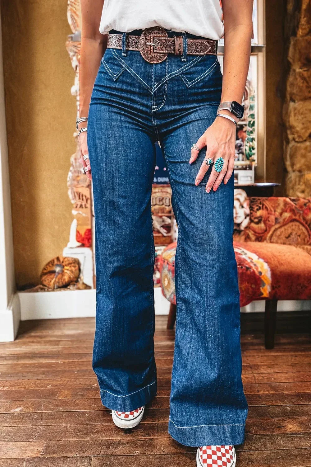 Woman wearing high-waisted flared blue jeans with white stitching, a brown embossed leather belt, and red checkered sneakers