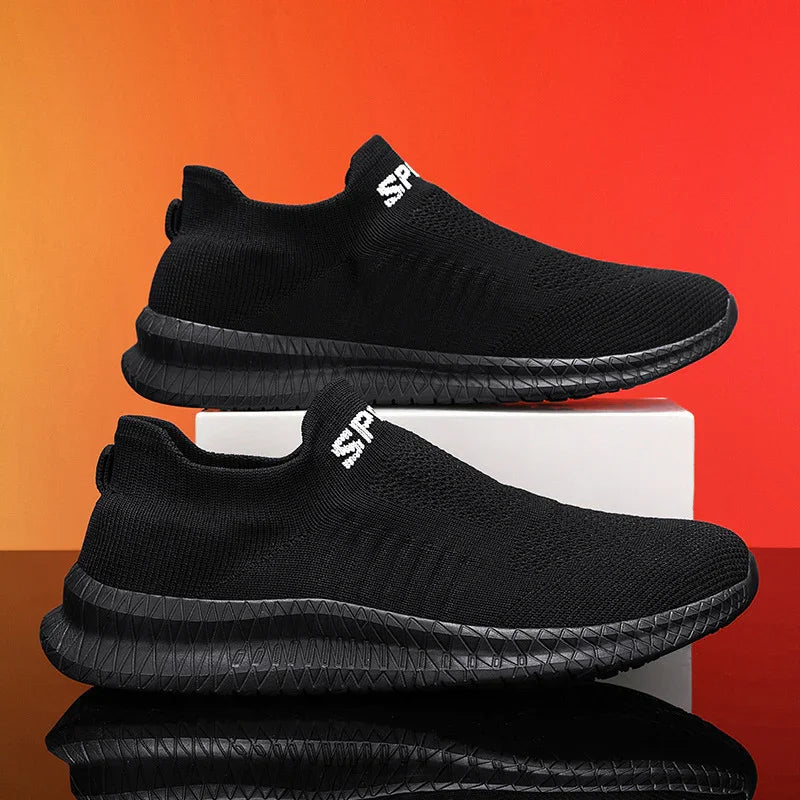 Pair of black slip-on sports shoes with textured black soles displayed on white box against orange background